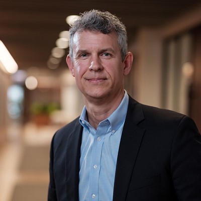 Portrait of a middle-aged man with gray curly hair wearing a light blue button-up shirt and a black blazer, standing in a softly lit indoor hallway with blurred background.