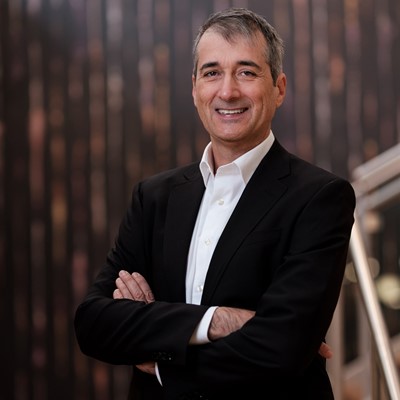 A middle-aged man with short gray hair wearing a black suit jacket and white dress shirt stands with his arms crossed, smiling confidently against a blurred dark brown and black background.