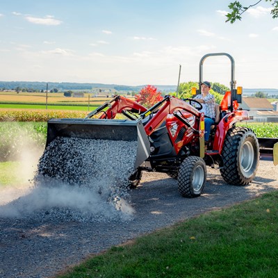 A person operating a red tractor with a front loader, spreading gravel on a rural dirt road. The background features green fields, a farmhouse, silos, and a clear blue sky with scattered clouds.