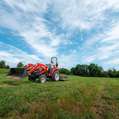 A man driving a red tractor with a front loader attachment across a green grassy field under a partly cloudy blue sky, with trees in the background.