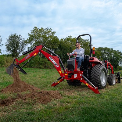 A man operating a red CASE IH tractor equipped with a backhoe attachment, digging into the grassy ground in a rural outdoor setting with trees and a cloudy sky in the background.