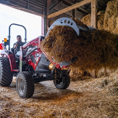 A person operating a red tractor inside a barn, using a front loader attachment to lift a large bale of hay. The barn is filled with stacked hay bales and straw scattered on the ground.