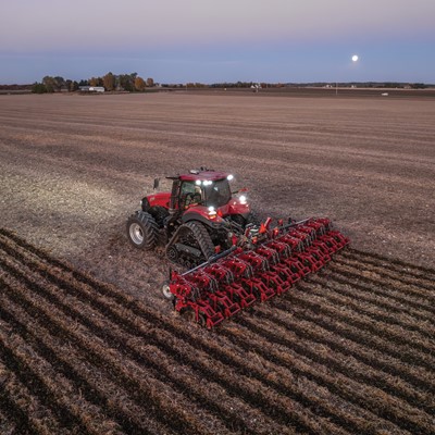 A red tractor with large tracks is plowing a vast, open field at dusk, creating evenly spaced furrows in the soil under a clear sky with the moon visible in the background.
