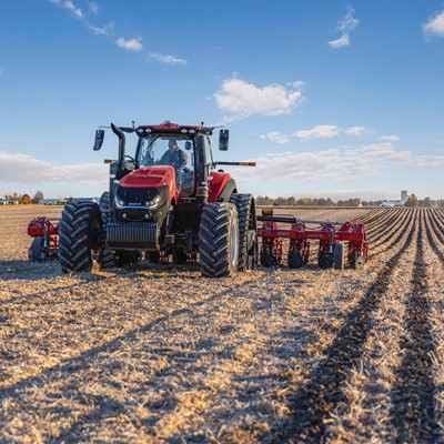 A red tractor with large tires is plowing a vast, dry agricultural field under a blue sky with scattered clouds. The tractor is positioned in the center of the image, moving towards the camera, creating evenly spaced furrows in the soil. Trees and farm buildings are visible in the distant background.