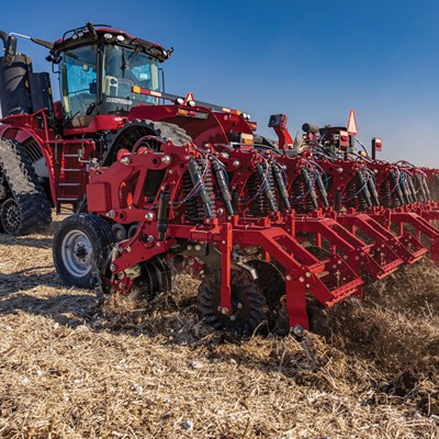 A large red agricultural tractor with rubber tracks is pulling a multi-row planter attachment across a dry field under a clear blue sky, preparing the soil for planting.