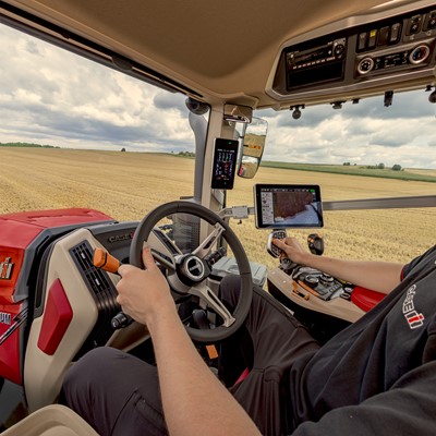 Interior view of a modern Case IH Optum tractor cabin showing a person operating the steering wheel and control panel, with a large golden wheat field visible through the windows under a cloudy sky.