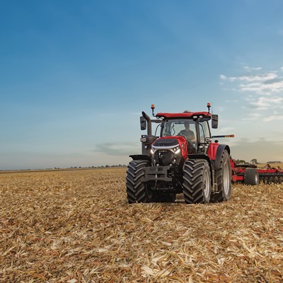 A red tractor with large tires is working in a vast harvested cornfield under a clear blue sky with scattered clouds during sunset.