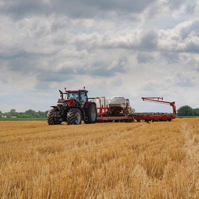 A red tractor with large tires is working in a golden wheat field under a cloudy sky, pulling farming equipment used for planting or fertilizing. Trees and greenery are visible in the distant background.