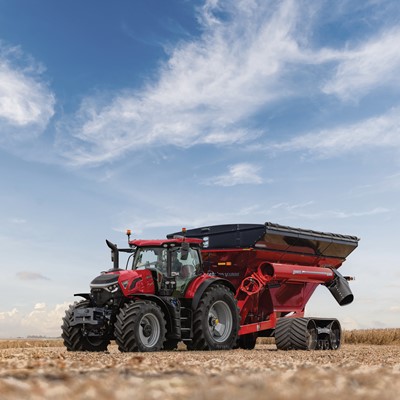 A red tractor attached to a large red grain cart with black tracks is parked in a harvested wheat field under a partly cloudy blue sky.