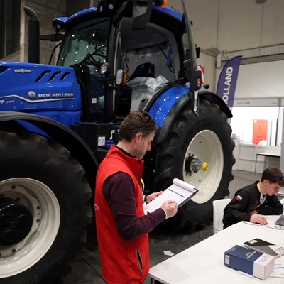 A large blue New Holland T7.300 tractor is displayed indoors at an exhibition or trade show. In the foreground, a man wearing a red vest is holding and writing on a clipboard, while another person is seated at a white table working on a laptop. Various banners and booths are visible in the background.