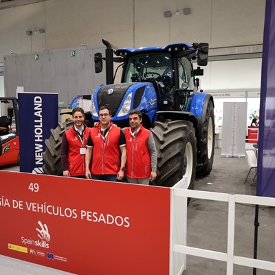 Three men wearing red vests standing in front of a large blue New Holland tractor at an indoor exhibition booth labeled "Tecnología de Vehículos Pesados" with SpainSkills branding.