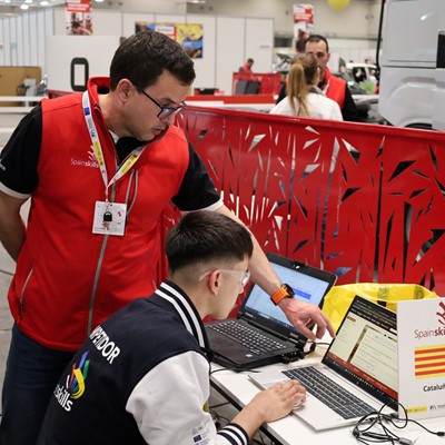 Two men working together on laptops at a SpainSkills event in an indoor workshop setting, with one man standing and pointing at the laptop screen while the other sits and types. A sign on the table displays the SpainSkills logo and the word "Cataluña" with the Catalonia flag. The background shows other participants and equipment in a spacious, well-lit area.