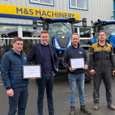 Four men standing in front of two blue tractors outside a building with a yellow and blue "M&S Machinery" sign. Two of the men are holding framed certificates, and all are dressed in casual or work attire. The scene appears to be an award presentation or recognition event.