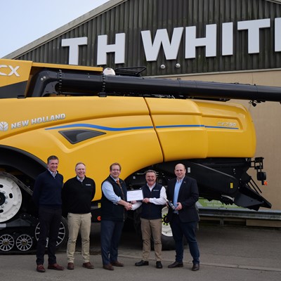 Five men standing in front of a large yellow New Holland CX combine harvester outside a building with a sign that reads "TH WHITE." Two of the men in the center are shaking hands and holding a certificate, while the others stand beside them smiling.