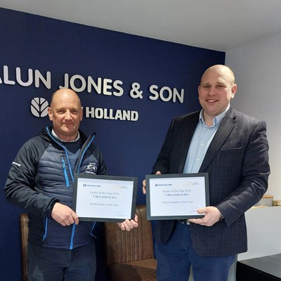 Two men standing indoors in front of a dark blue wall with the text "T. Alun Jones & Son" and "New Holland" logo. Both men are holding framed certificates. The man on the left is wearing a black jacket and dark pants, while the man on the right is dressed in a dark blazer, light blue shirt, and blue pants. There are two brown chairs against the wall behind them.