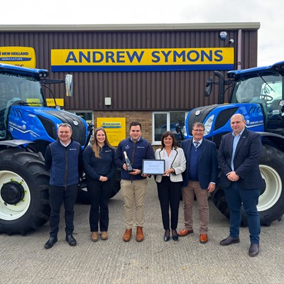 Six people standing in front of two large blue New Holland tractors outside a building with a yellow and blue "Andrew Symons" sign. One person in the center is holding a trophy, and another is holding a framed certificate, indicating an award or recognition event. The group is dressed in business casual and branded clothing.