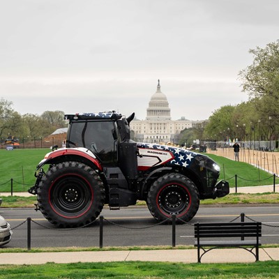 A large tractor painted with an American flag design is driving on a road near the National Mall in Washington, D.C., with the United States Capitol building visible in the background. There are a few parked cars, a bench, and trees lining the area under an overcast sky.