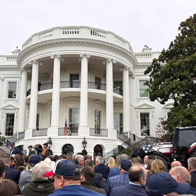 A large crowd of people gathered in front of the South Portico of the White House, with a tractor decorated in red, white, and blue parked to the right side near a large tree, under an overcast sky.