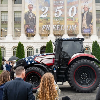 A large tractor painted with an American flag design is displayed outdoors in front of a historic building with tall columns. A banner hanging on the building reads "USDA Celebrates 250 Years of Freedom" with images of Abraham Lincoln and George Washington. Several people stand nearby, some taking photos, observing the tractor and the event. The scene appears to be a public celebration or exhibition.