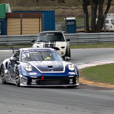 Two race cars navigating a curved section of a racetrack, with a blue and white Porsche leading and a white Mini Cooper with black racing stripes following closely behind, surrounded by safety barriers and grassy areas.