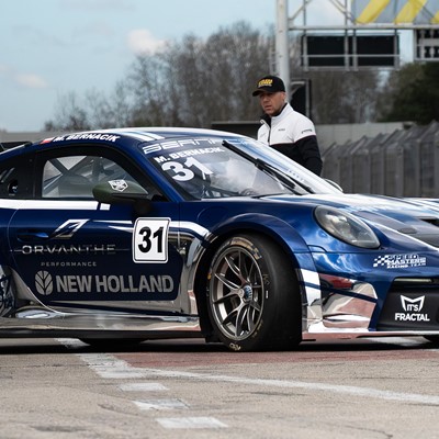 A blue and white race car with the number 31 on the door is parked on a race track. The car features sponsor logos including New Holland, Orvanthe Performance, and ITS Fractal. A man wearing a black and white jacket and a black cap stands behind the car, near the track barrier. The background shows a cloudy sky and some trees.