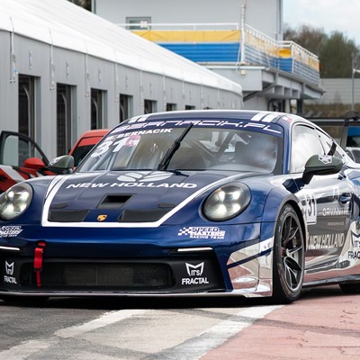 A blue and white Porsche race car with the number 31 and "New Holland" branding is parked on a race track pit lane next to a row of garages. The car features racing decals, a large rear wing, and a red tow hook on the front bumper. The background shows a partly cloudy sky and some greenery.