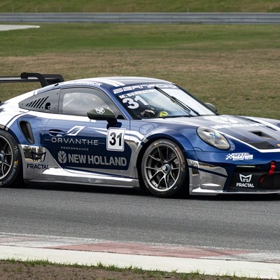 A blue and silver Porsche race car with number 31 on the door is driving on a race track. The car features sponsor logos including Orvanthe Performance, New Holland, XPEL, and Fractal, and has a large rear wing for aerodynamics. The background shows grassy areas and tire barriers along the track.