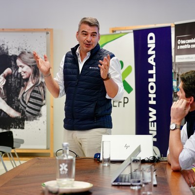 Two men in a meeting room, one standing and speaking with expressive hand gestures while the other sits at a table listening attentively. Both are wearing navy blue vests over white shirts. The table has laptops, glasses of water, and a water bottle. In the background, there are banners with the logos "NEW HOLLAND" and "RIGOR," and a poster featuring a woman working with her hands. The setting appears to be a professional or corporate environment.
