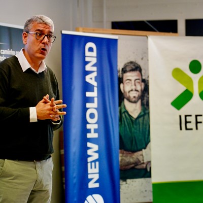 A man with gray hair and glasses, wearing a black sweater over a white collared shirt and beige pants, is speaking or presenting indoors. Behind him are three vertical banners, one blue with the text "NEW HOLLAND," another with a photo of a man with crossed arms, and a white banner with a green and yellow logo and the text "IEFP.