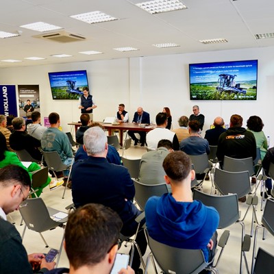 A group of people attending a presentation or seminar in a conference room. A speaker stands at the front near two large screens displaying slides with agricultural machinery images and text. Several panelists are seated at a table beside the speaker. Attendees are seated in rows facing the front, some taking notes or using their phones. Banners for IEFP and New Holland are visible in the background. The setting appears professional and focused on agricultural or farming topics.