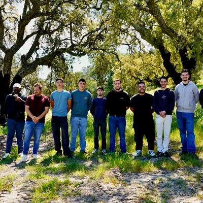 A group of ten young men standing outdoors in a sunny, grassy area with large trees in the background, all facing the camera and posing for a group photo.