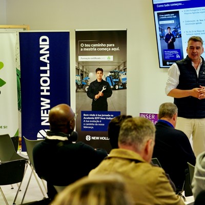 A man in a white shirt and black vest is giving a presentation to an audience seated in a room. Behind him are banners for IEFP and New Holland, and a screen displaying information in Portuguese about a training course and collaboration between IEFP and New Holland. The audience is attentively listening, and the setting appears to be a professional or educational event focused on agricultural machinery training.
