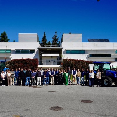 A large group of people standing in front of a modern white building with green bushes and trees in the background. Two blue New Holland tractors are positioned on either side of the group, with New Holland banners next to each tractor. The sky is clear and blue, indicating a sunny day.