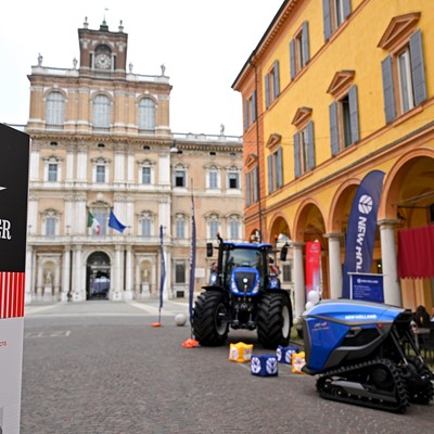 Outdoor exhibition of New Holland agricultural machinery including a blue tractor and a tracked vehicle displayed on a cobblestone street in front of historic buildings with arches and a clock tower in the background, during the Motor Valley Fest in Emilia Romagna, Italy.