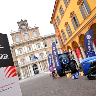 Outdoor event setup in a historic Italian square featuring a Motor Valley Fest sign with logos of Visit Emilia Romagna and RER, alongside New Holland branded agricultural machinery including a blue tractor and a blue tracked vehicle, with a person on a bicycle nearby and classical architecture in the background.