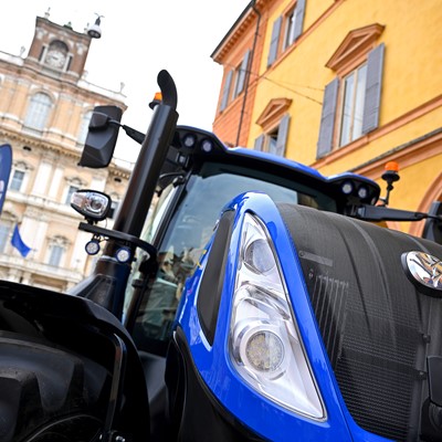 Close-up view of a blue New Holland tractor displayed outdoors in front of historic European-style buildings with arched windows and shutters, under a partly cloudy sky.