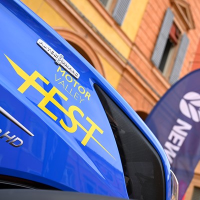 Close-up of a blue vehicle with yellow text "Motor Valley Fest" and model designation "T7.340 HD" on its side, set against a background of colorful buildings and a blurred flag with the word "New" visible.