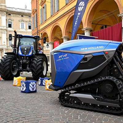 A blue New Holland R4 Hybrid Power autonomous agricultural vehicle with tracks is displayed on a cobblestone street next to a traditional blue New Holland tractor with large tires. The scene is set in an urban area with historic buildings and arches in the background, accompanied by New Holland branded flags and colorful cube seats on the ground.