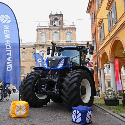 A large blue New Holland tractor is displayed on a cobblestone street in front of historic buildings with arched windows and a clock tower. Blue New Holland flags and branded cube cushions are placed around the tractor. People walk and ride bicycles nearby, enjoying the outdoor event.