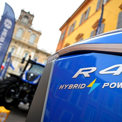 Close-up view of the front side of a blue R4 Hybrid Power tractor with a blurred background showing another tractor, flags, and historic buildings in an outdoor urban setting.