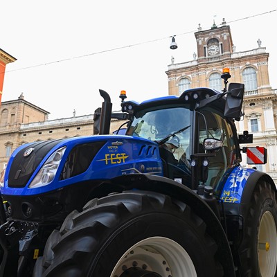Blue New Holland tractor with "Motor Valley Fest" branding displayed in an urban setting with historic buildings in the background, under a clear sky.
