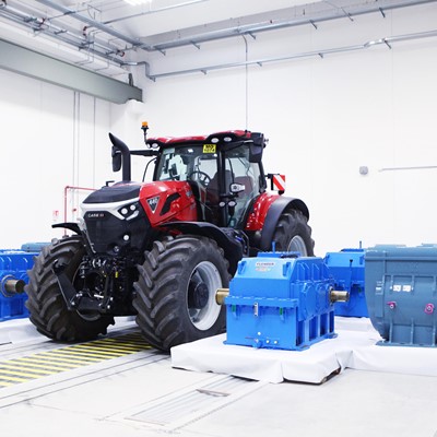 A red Case IH tractor is positioned indoors on a testing platform surrounded by several large blue industrial gearboxes and machinery components, all set in a clean, well-lit white room with visible ceiling pipes and safety markings on the floor.