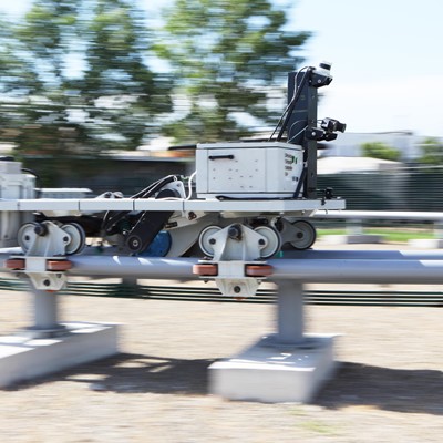 A mobile robot equipped with cameras and sensors is moving along a metal rail outdoors, with blurred trees and buildings in the background indicating motion.
