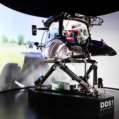 A high-tech driving simulator setup featuring a tractor cabin mounted on a multi-axis motion platform labeled "DDS1," with a large curved screen in the background displaying a virtual outdoor environment, designed for immersive agricultural machinery training or testing.