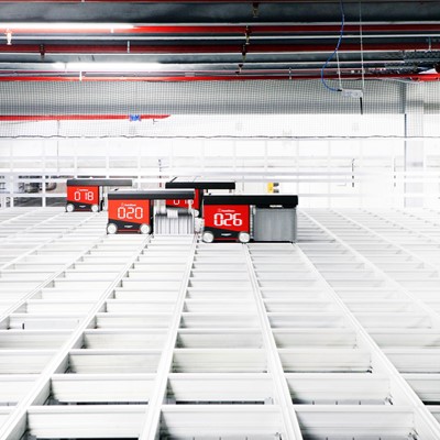 A high-tech automated warehouse with red robotic carts numbered 018, 020, and 026 moving along white grid-like storage shelves under a ceiling with red pipes and industrial lighting.