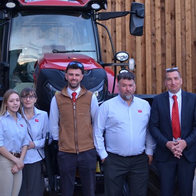 A group of eight professionally dressed people standing in front of a large red tractor and a wooden wall, posing for a group photo. Most individuals are wearing light blue shirts, some with red ties, and one person is wearing a brown vest.