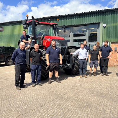 A group of nine men standing and posing in front of a large red tractor outside a green industrial building with the sign "Lloyd" on it. One man is sitting on the tractor, while the others stand on the ground, smiling and dressed in casual work attire. The scene is set on a sunny day with a blue sky and some clouds.