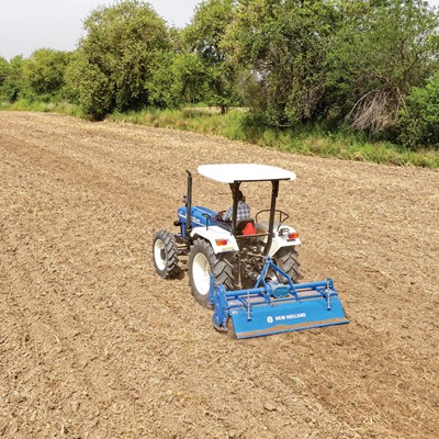 A blue and white New Holland tractor is tilling a large, dry, and freshly plowed field surrounded by green trees under a clear sky.