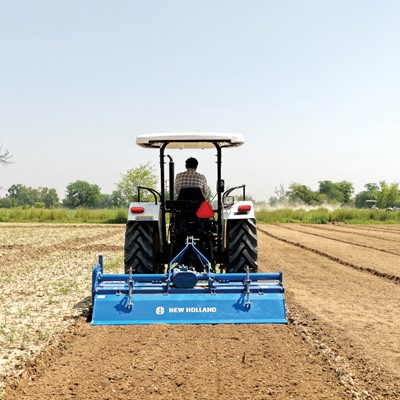 A farmer driving a white New Holland tractor with a blue tiller attachment, plowing a large, dry agricultural field under a clear sky, with green trees and other tractors visible in the background.