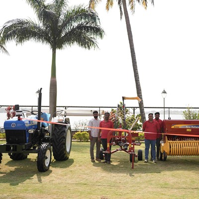A group of six men standing outdoors on grass near agricultural machinery, including a blue tractor on the left and a large red New Holland hay baler on the right, with palm trees and a body of water in the background under a clear sky.